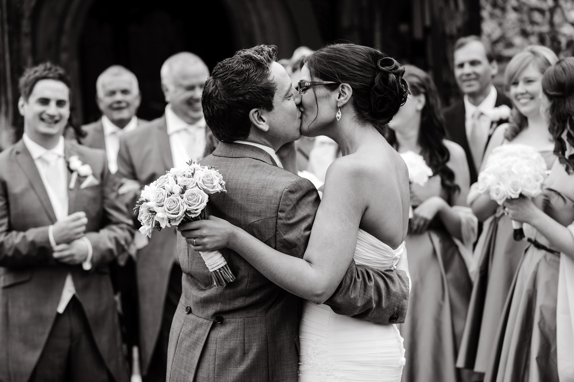 A photograph of a newly married couple kissing in front of their family and friends after their wedding ceremony taken by wedding photographer Richard Eaton.